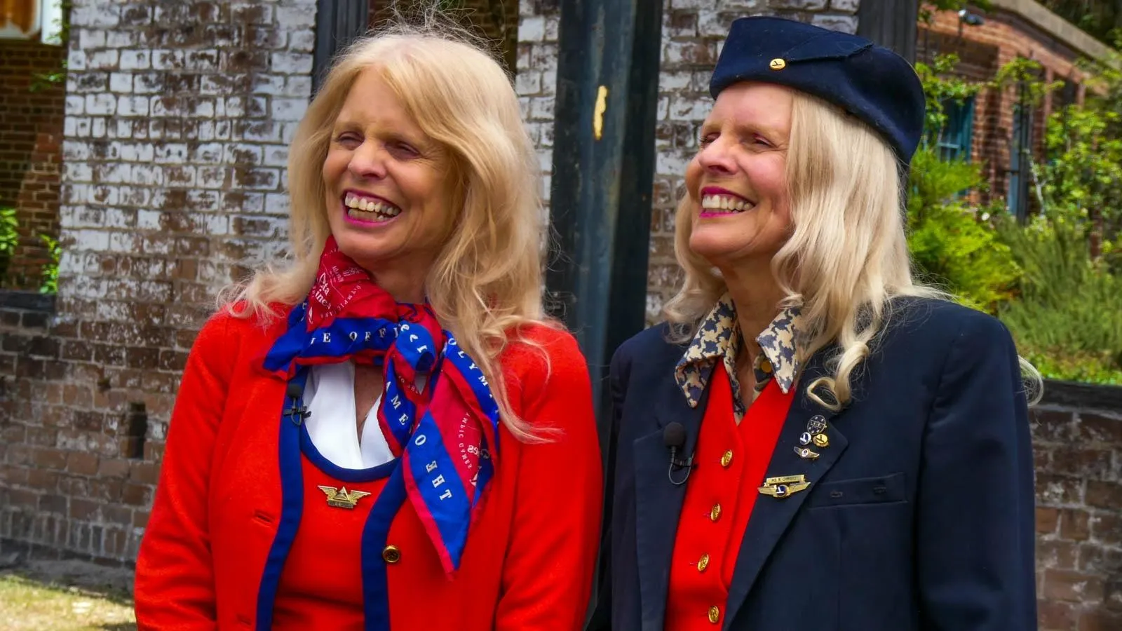 Two blonde women in flight attendent uniforms. The woman on the left is wearing a red uniform with blue accents, and the one on the right is wearing a blue uniform with a red under layer.