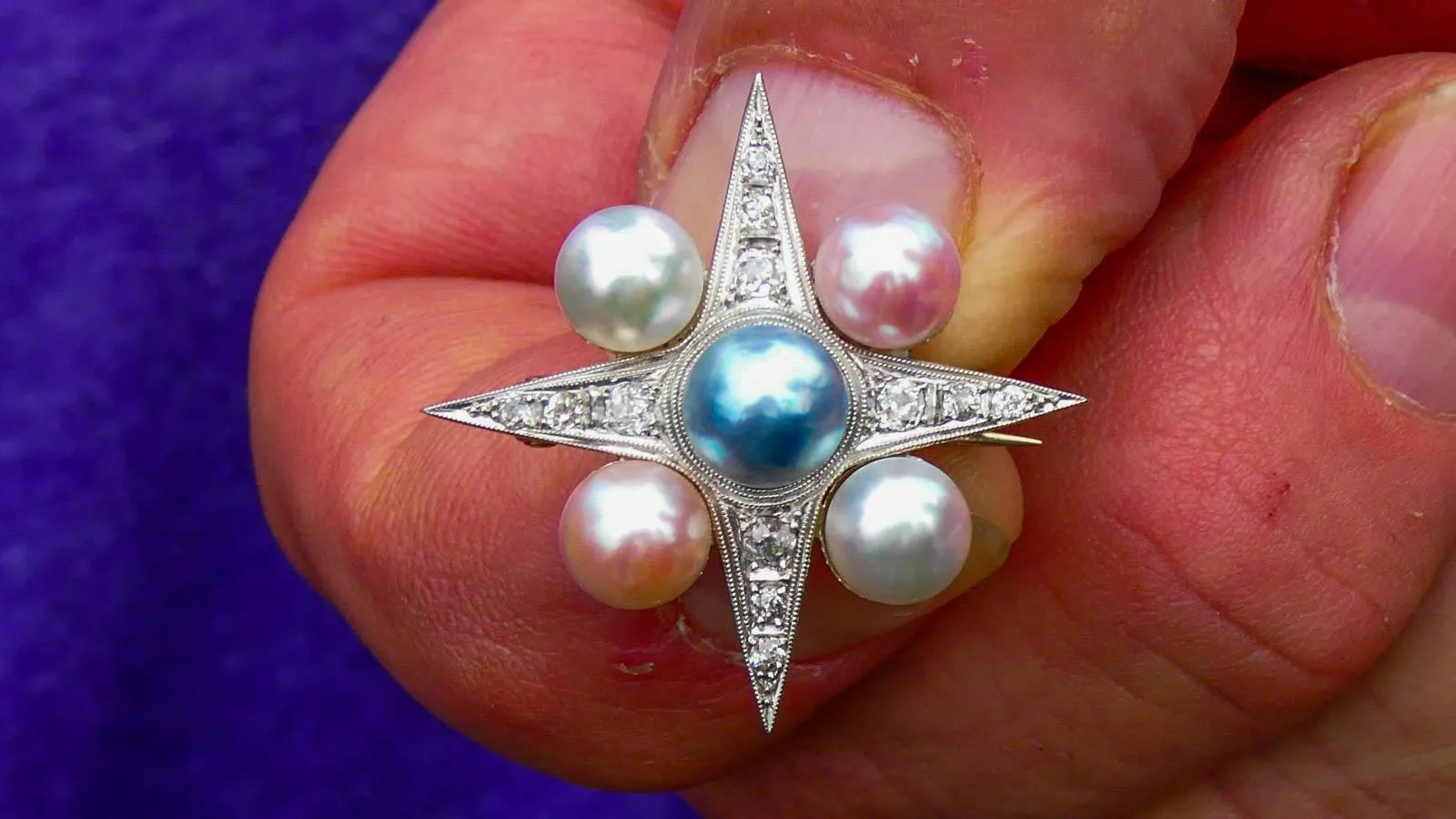 Between two pinched fingers sits a diamond four-point star with pink and blue pearls set in each corner with a darker blue pearl in the center.