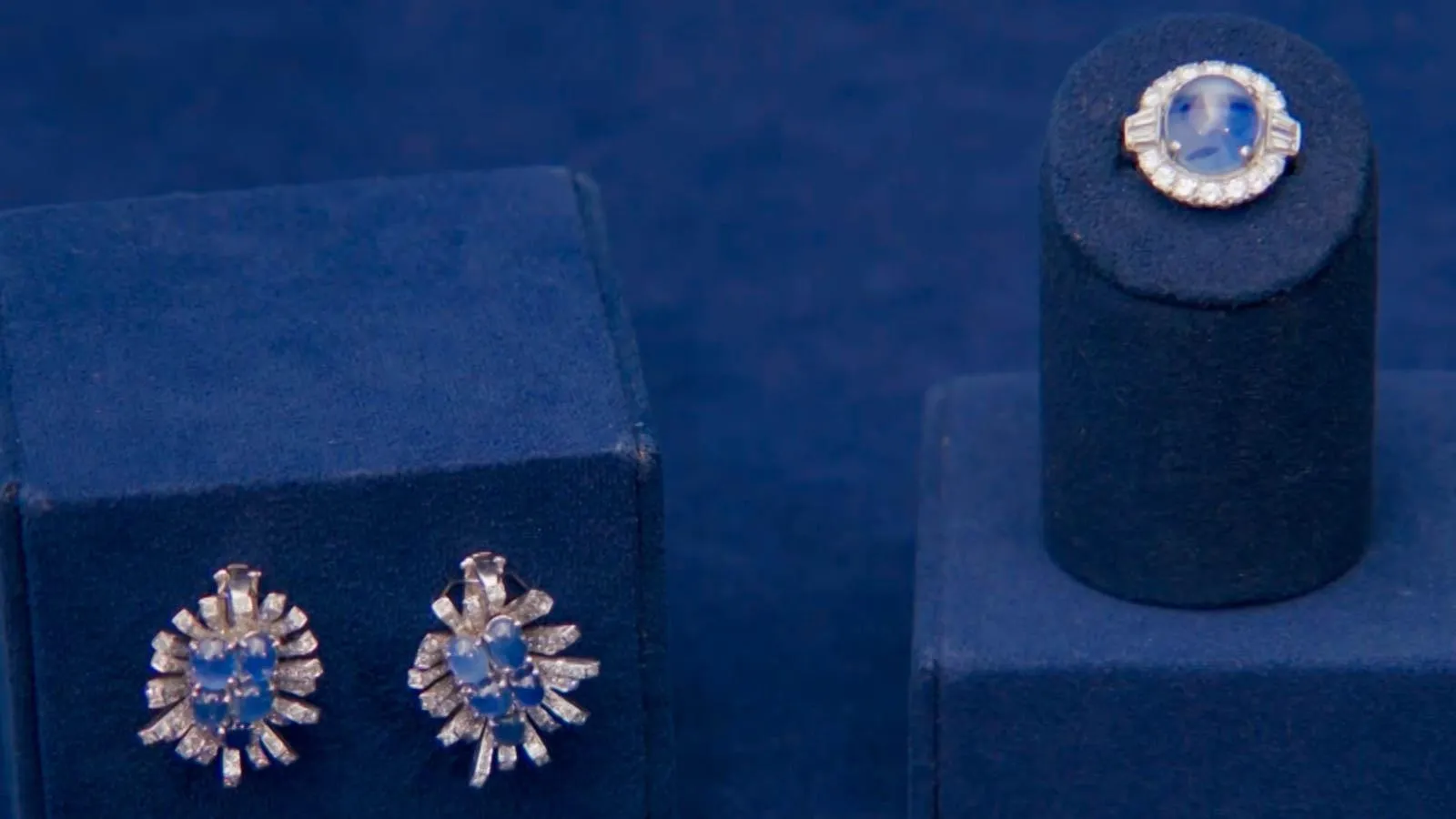 A pair of sapphire and diamond earnings next to a matching ring  mounted on a navy blue jewelry stands.
