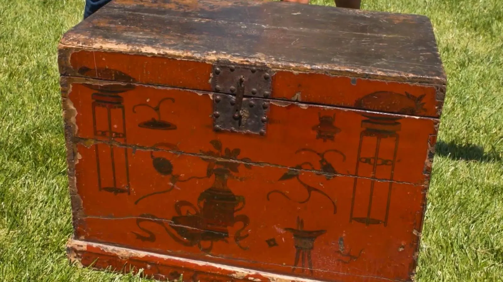 Close up of a large chest sitting on the grass. The chess has chipped reddish-brown paint on the front of it.  In dark brown paint, we see various Chinese art like vases and flowers.