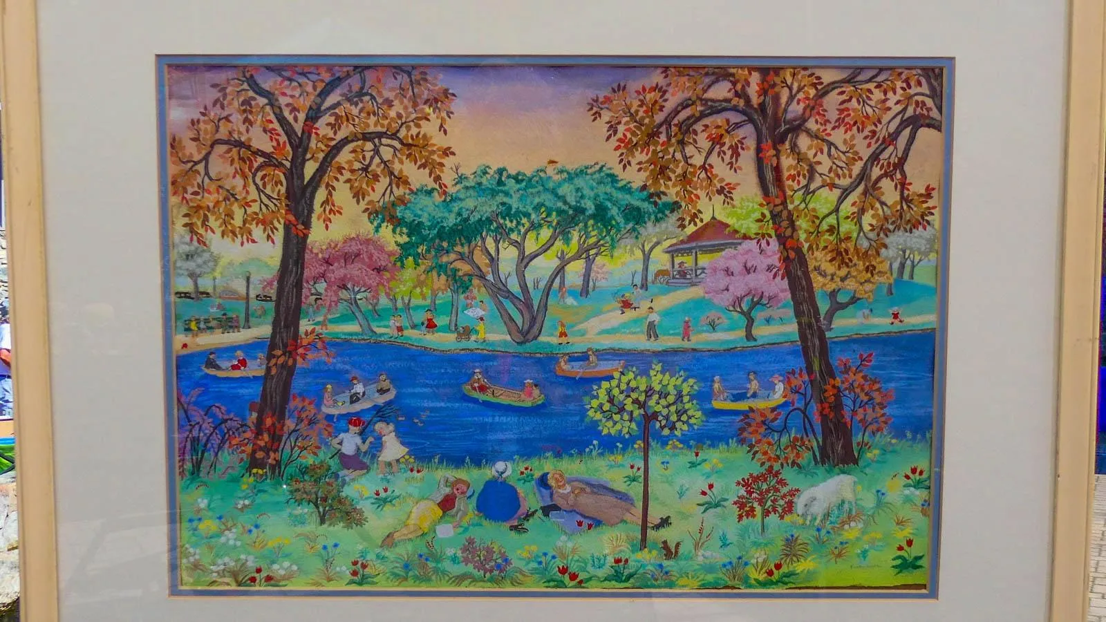 A vibrant watercolor in a wooden frame depicting people hanging out in a park near a river.