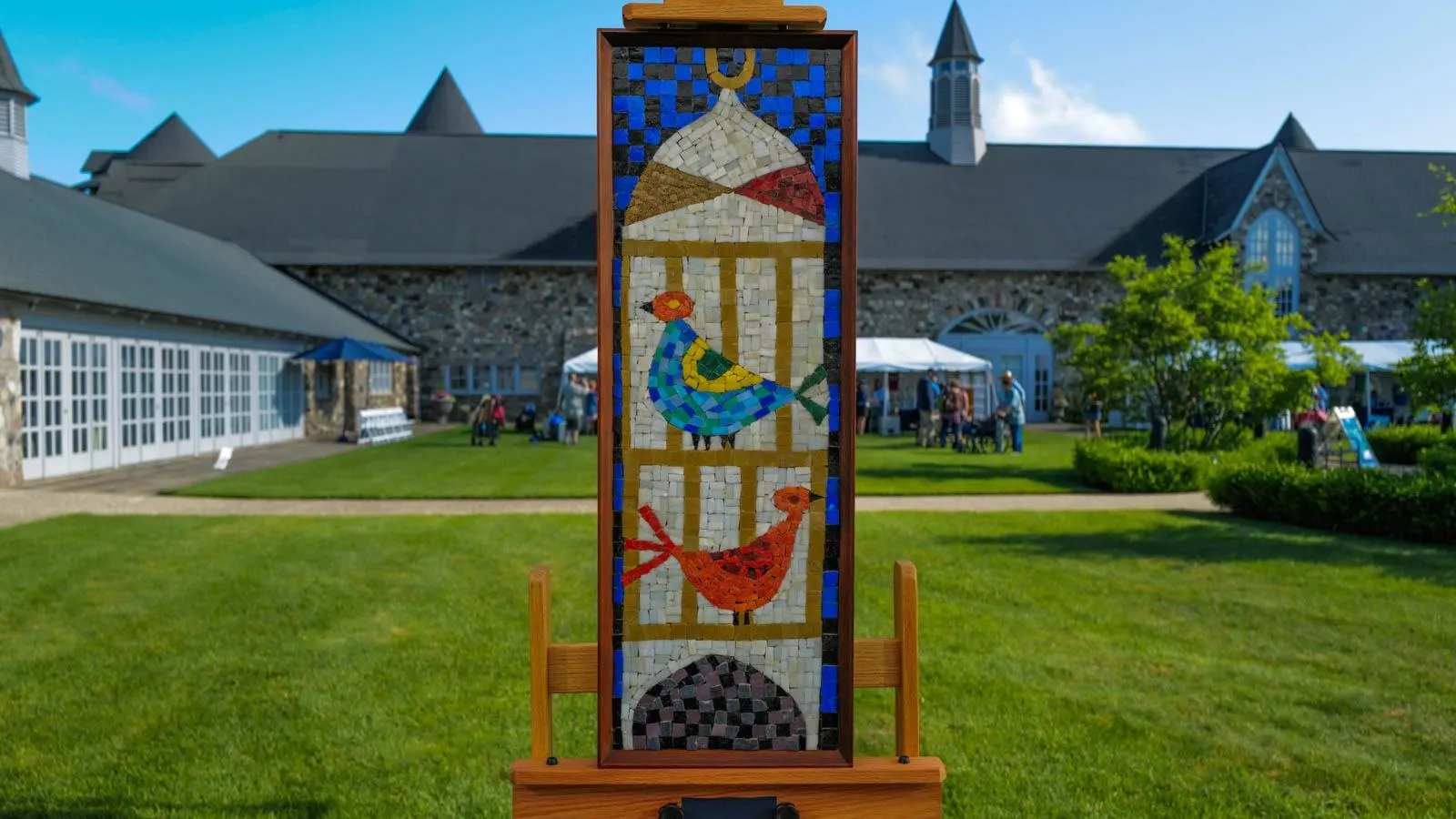 A mounted mosaic of two birds in a cage against an easel against and outdoor backdrop.