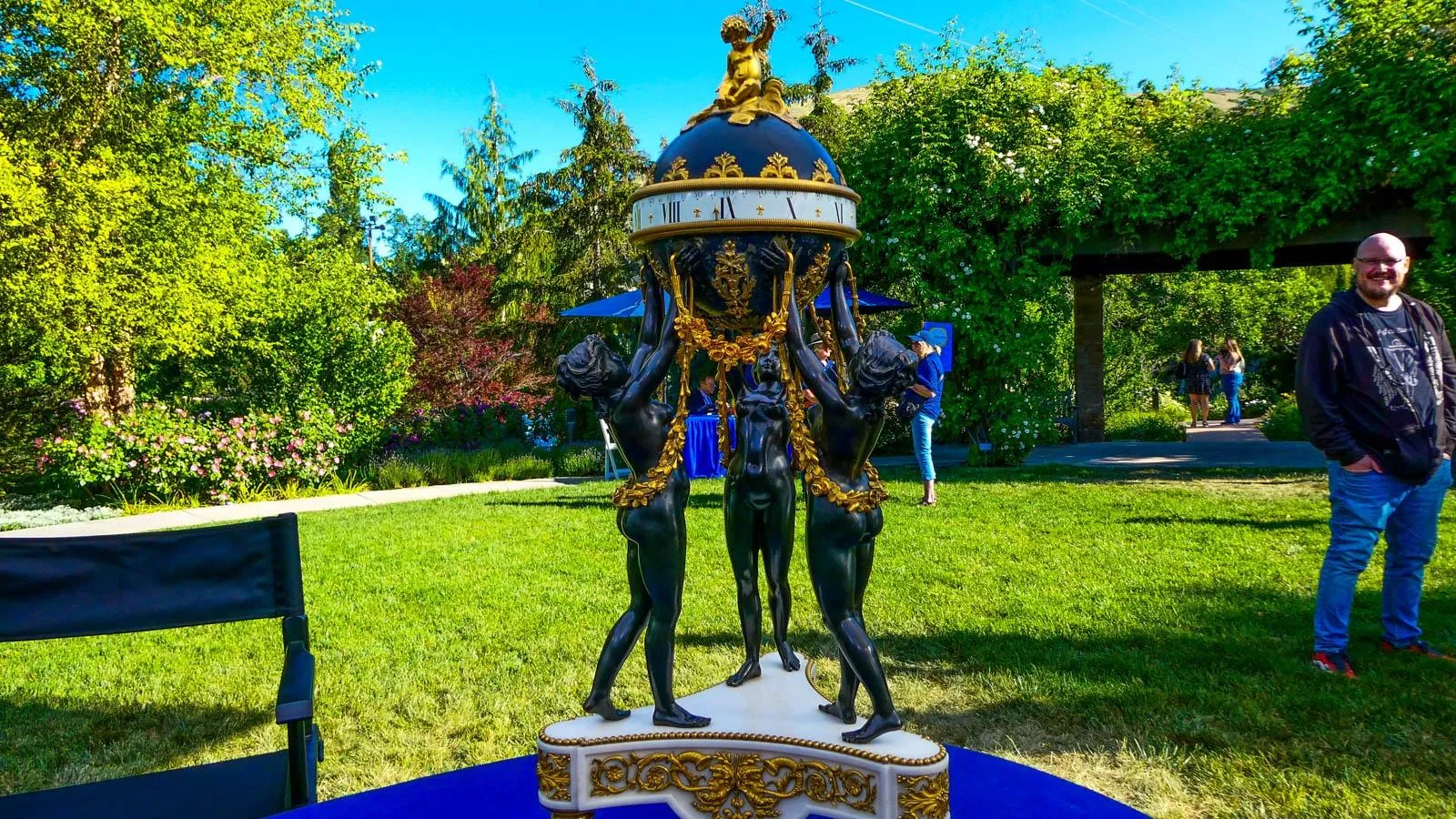 Three statue like figures uphold a globe with roman numerals around its circumference with gilded detaiks on the figures and base.