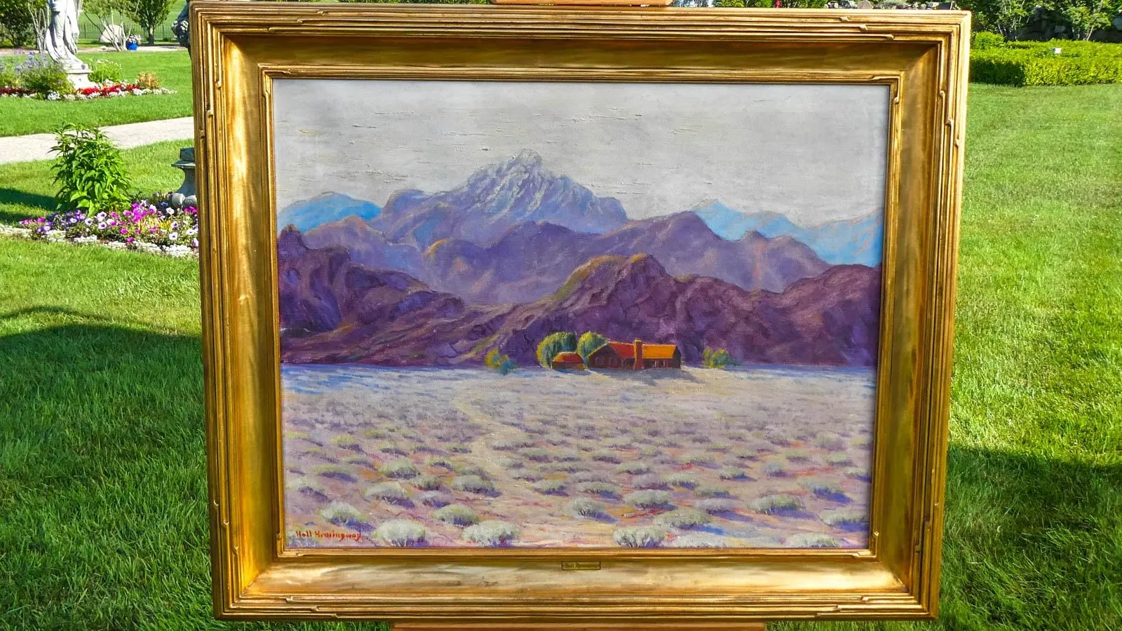 An oil painting of a desert mountainous landscape with purple hues placed in a golden frame mounted on an easel outdoors.