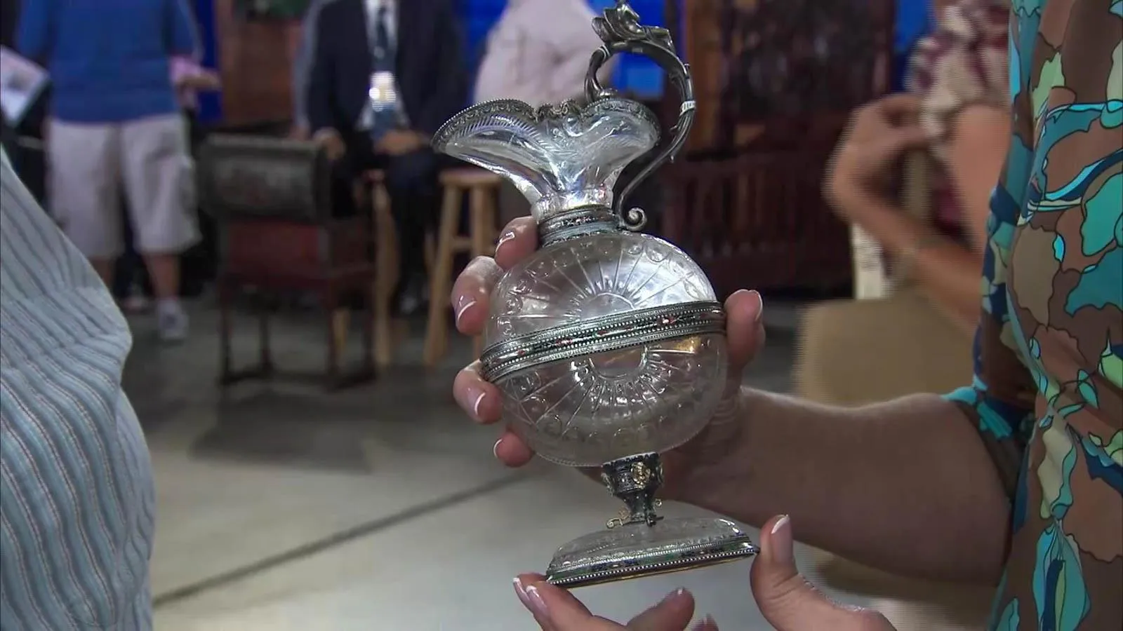 Close up of a silver Urn with a glass base, body and neck. The handle is silver and the piece has a silver rim on the bottom and top of the urn's spout.