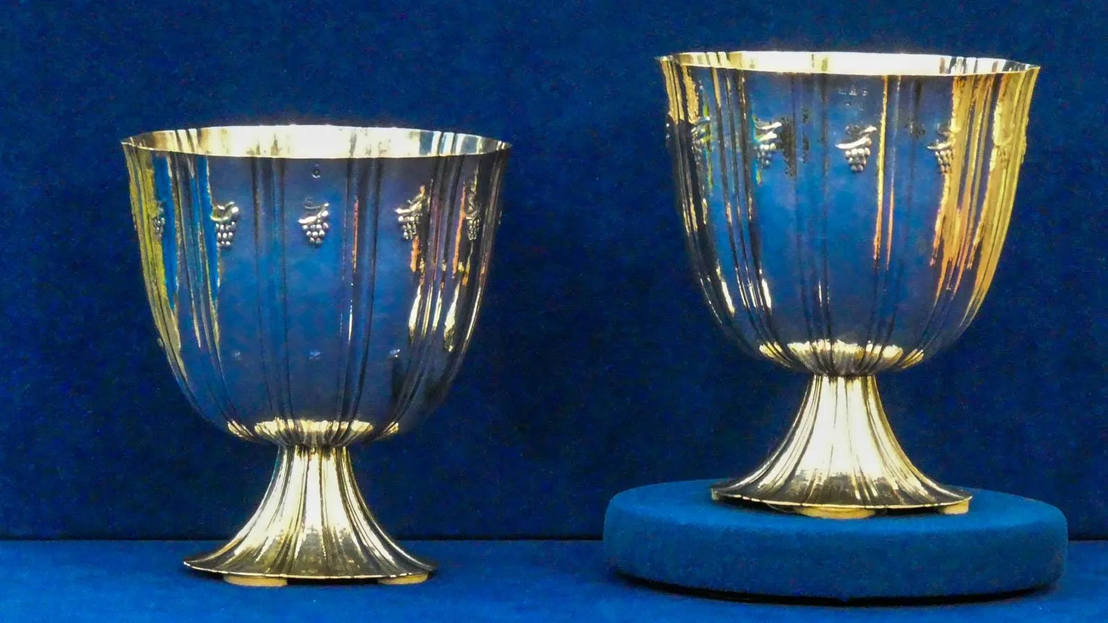 Two silver cups sit side by side against a blue backdrop. Across the rim of the cup is tiny delicate grape details.