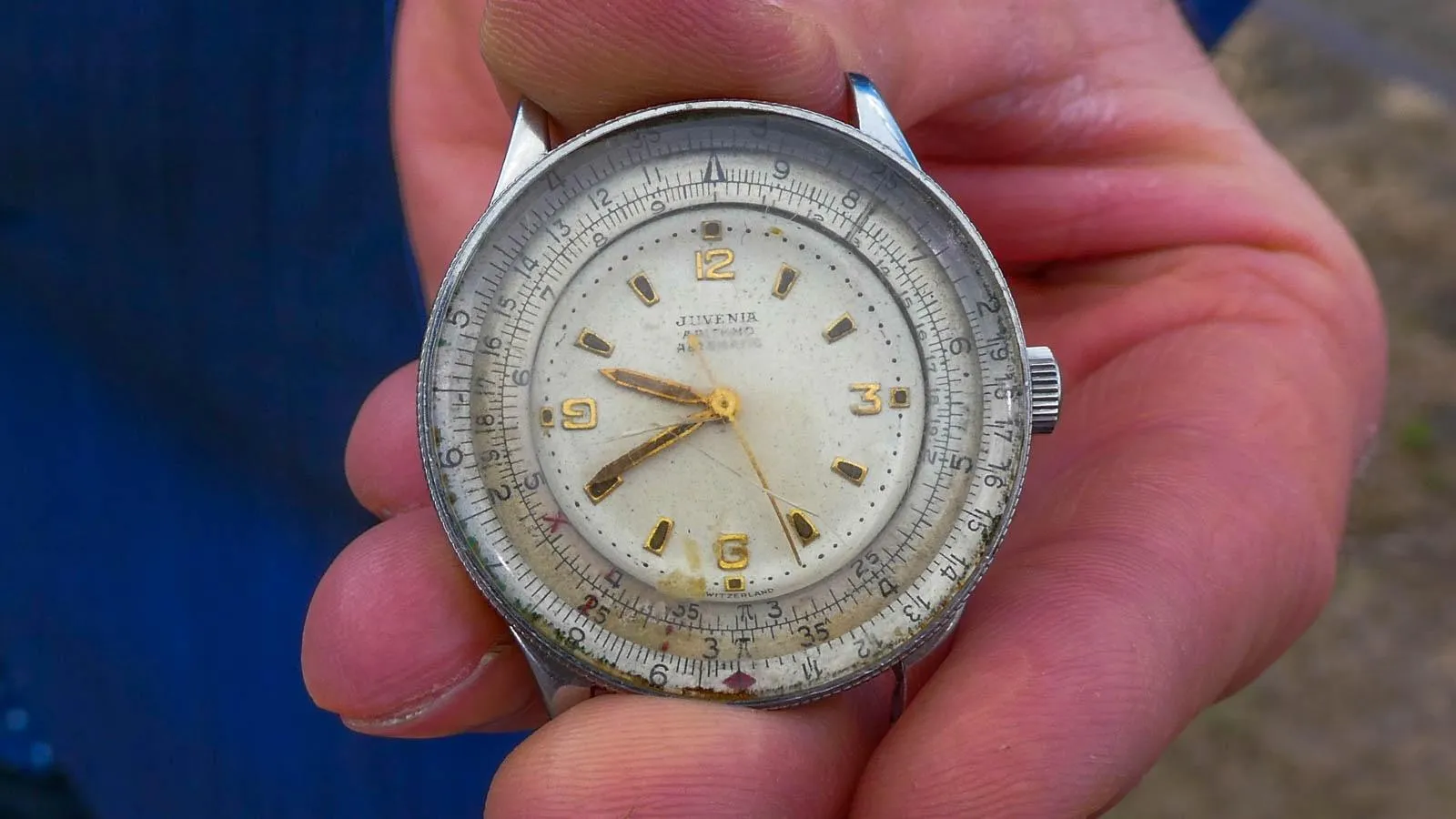 A hand is holding a round silver watch with black numbers around the rim and white watch face with gold numbering in the center.