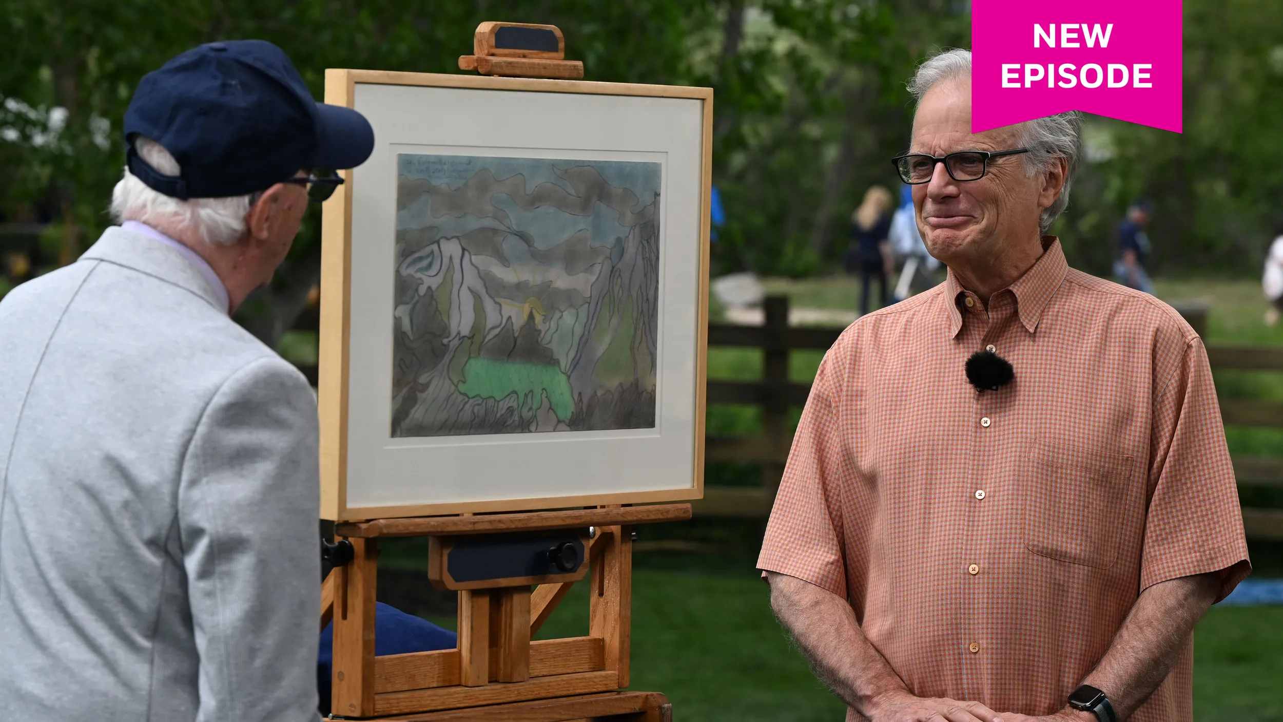 An older man in a red button down shirt looks at an appraiser, whose back is to the camera. The man looks tearful yet joyful. In between them sits a painting of the Italian Dolomites mountain range.