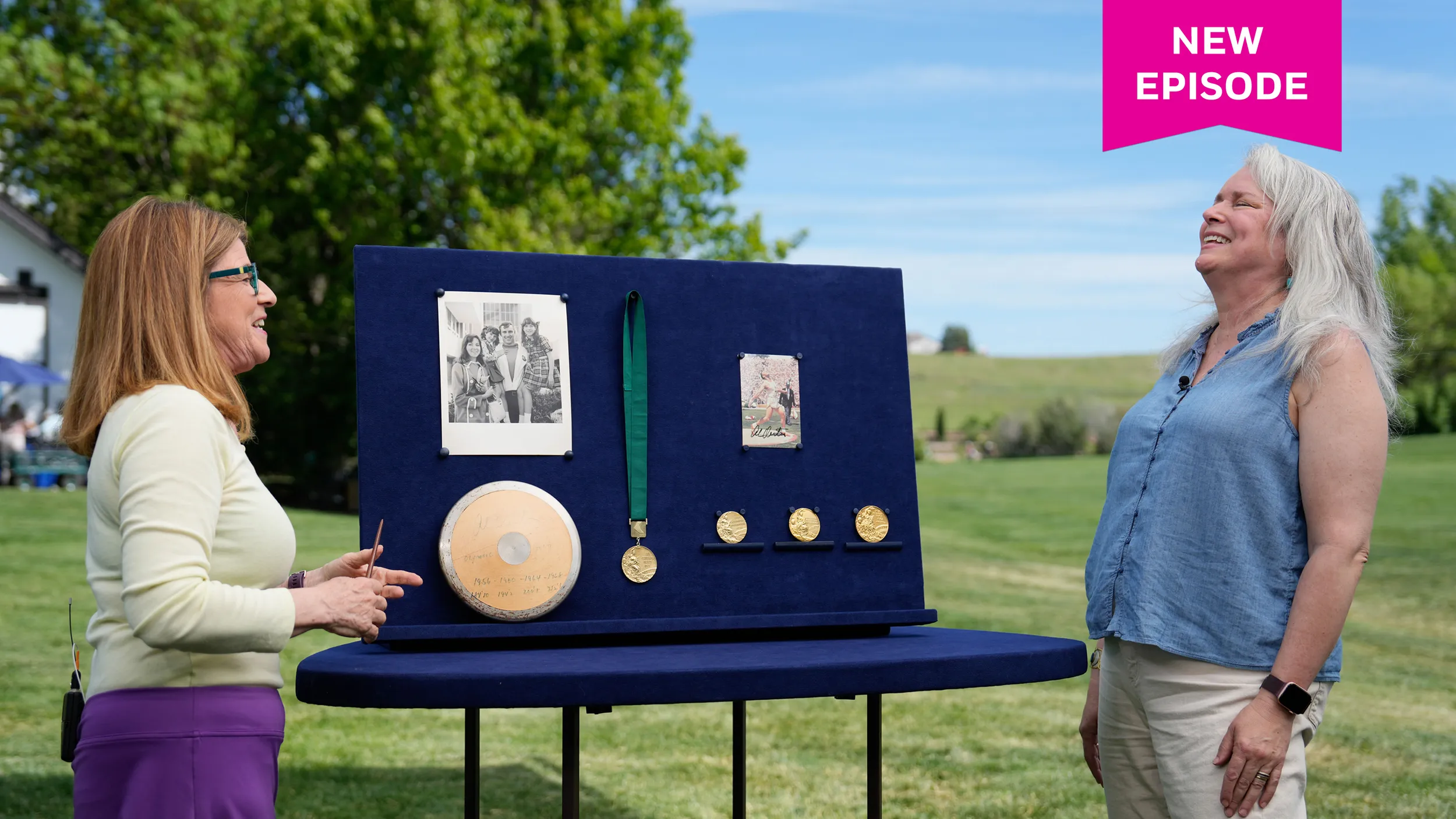 A female guest with white hair closes her eyes, leans back and laughs in delight at something the appraiser said. In between them is a collection of Olympic gold medals, a shot put, and photographs.