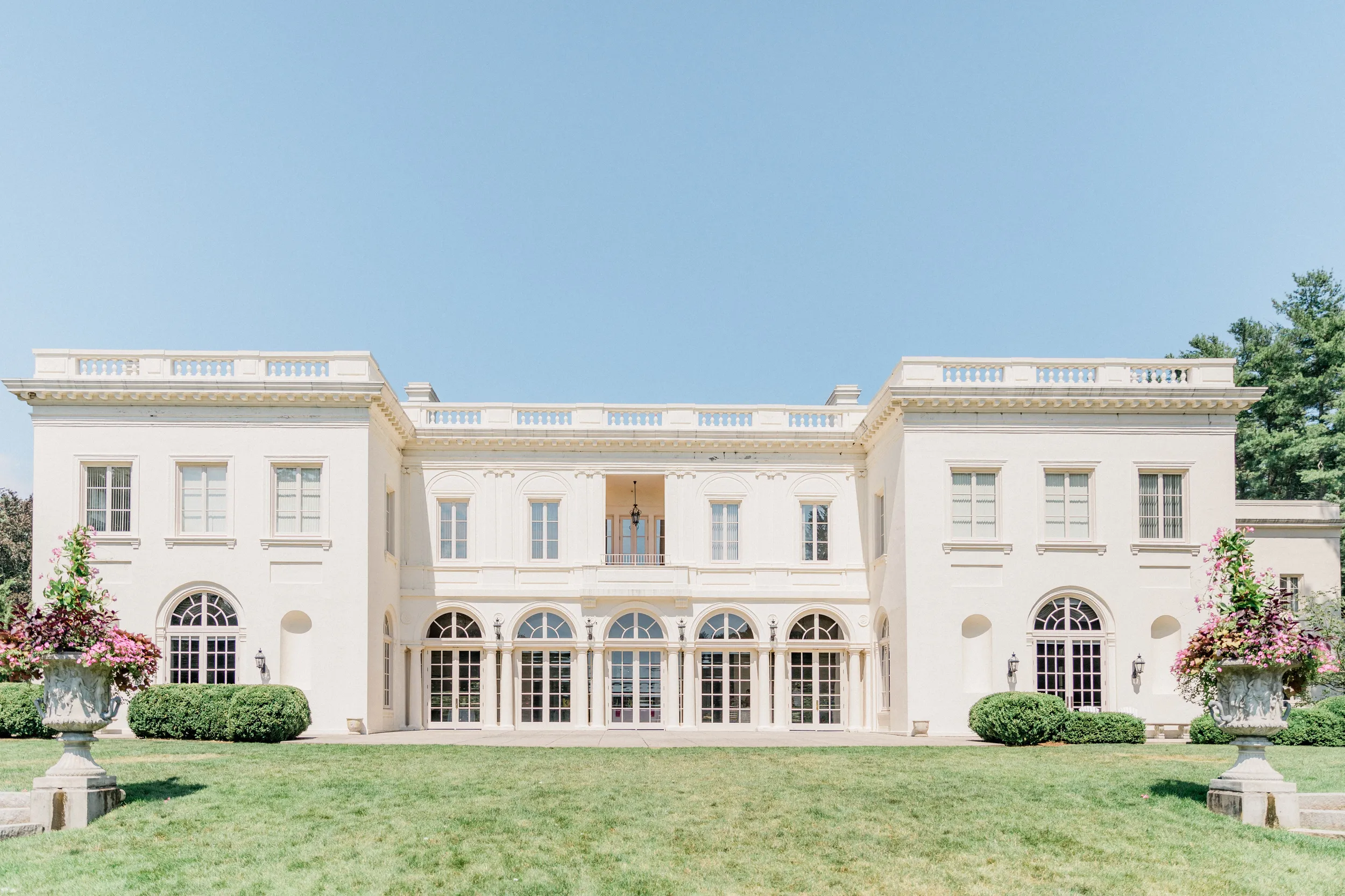 Exterior shot of a white two-story mansion that has a terrace. Front yard is visible, includes green bushes and two large sculptural planters with flowers.
