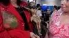 A woman in a red Blazer adorned with rhinestones and patchwork holds a crucifix with a sculpture of Jesus Christ covered in blood is affixed. To the right of the crucifix is a woman looking at it.