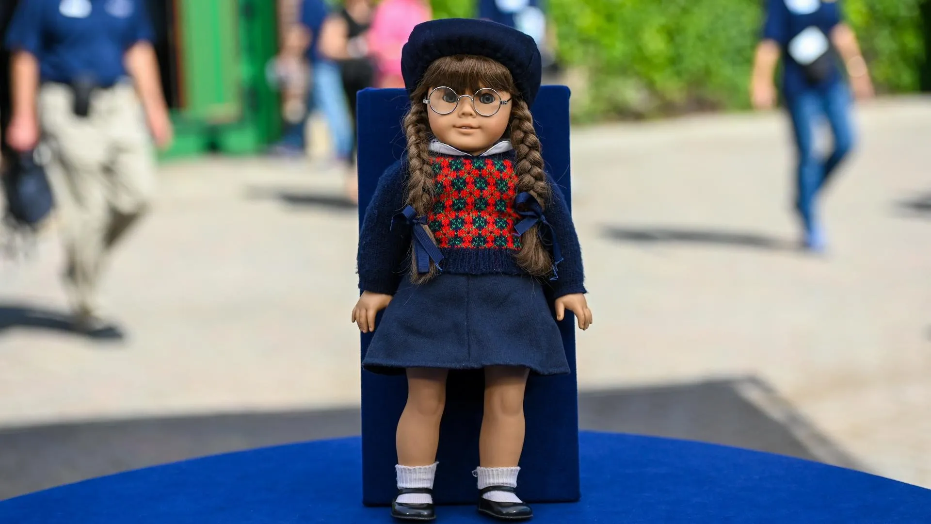 A doll in a plaid sweater, glasses, baret, and braids is placed on a blue display board on a blue table.