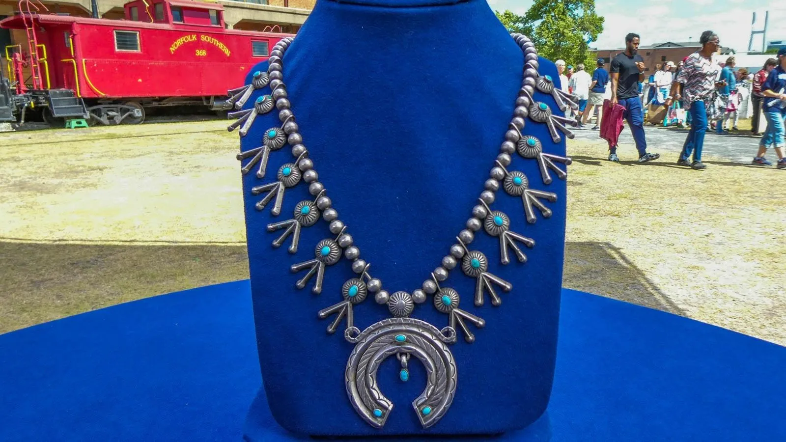Placed on a blue Jewlery holder sits a necklace of various round beads and accents of turquoise. In the center is a bigger medallian resembling a horseshoe with turquoise accents as well.