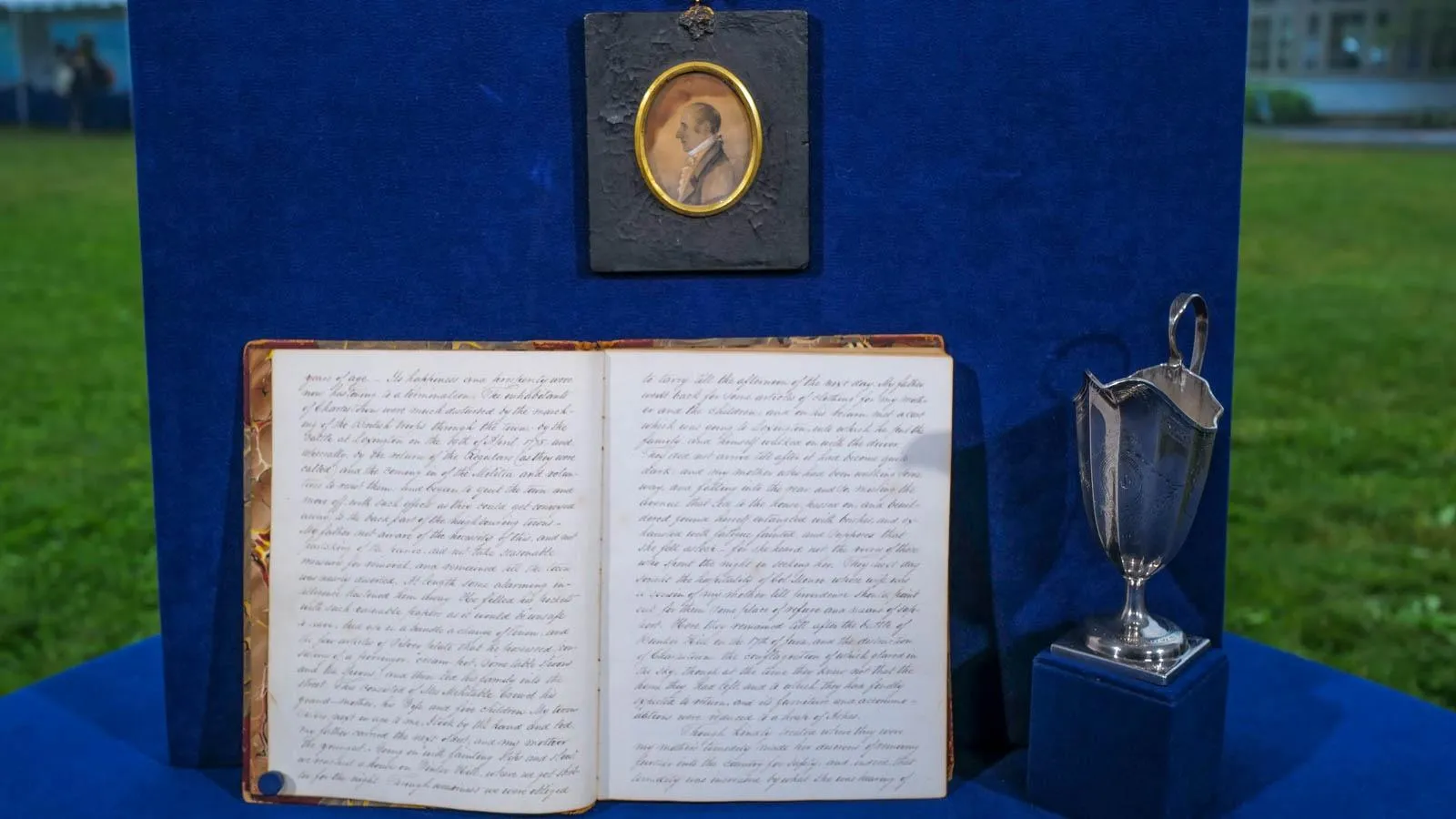 A diary written in cursive script is placed next to a small silver pitcher on a blue table just below a small oval portrait of a man.