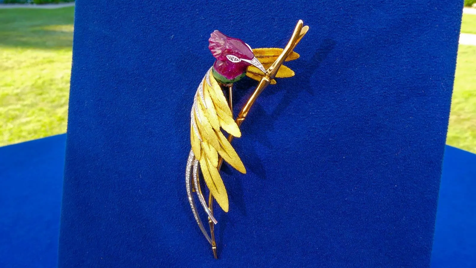 A bird brooch with a purple head and gold body with rhinestone accents mounted to a blue display board.