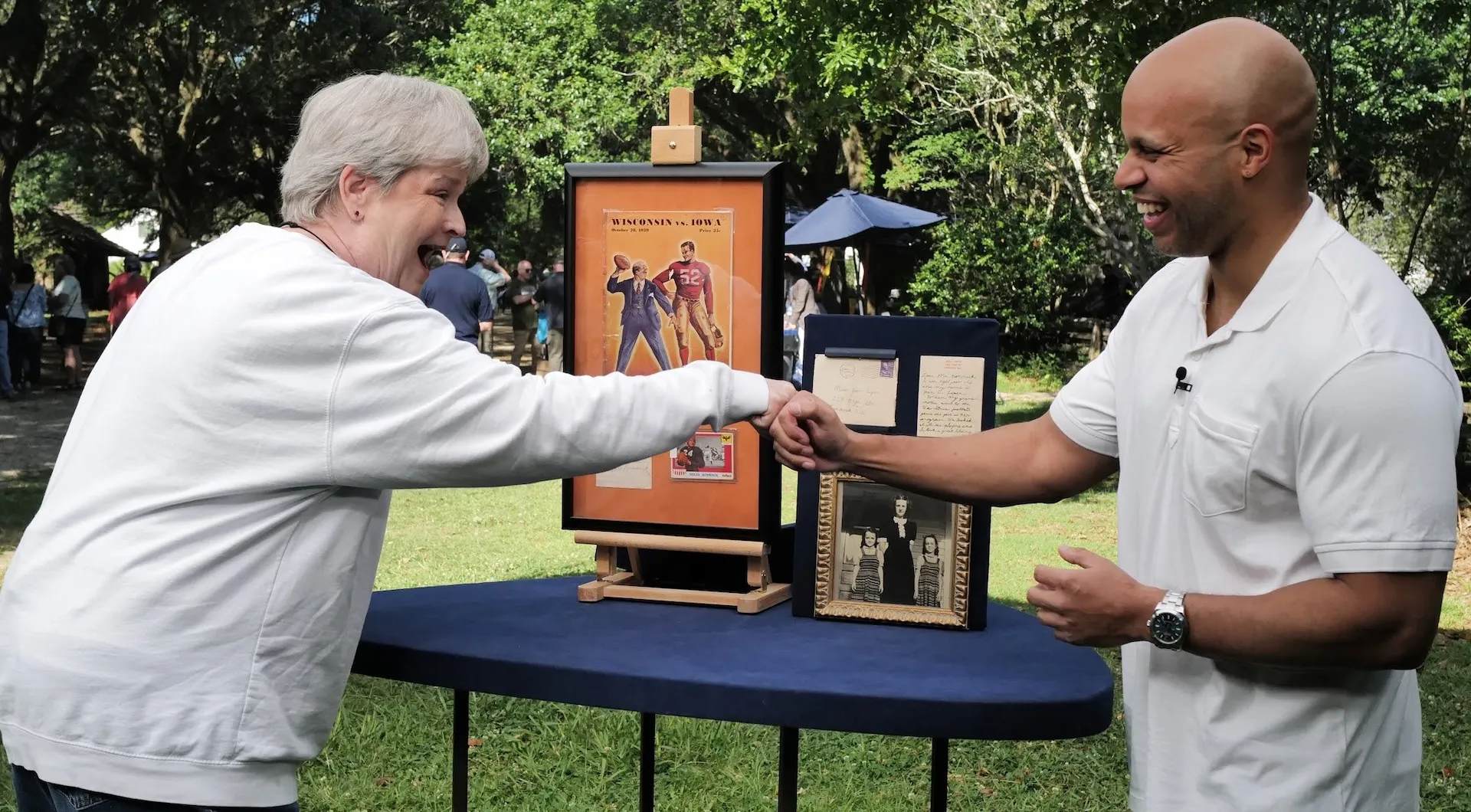 Photo of Sports Memorabilia appraiser Jasmani Francis (right) fist-bumping a female guest (left), both smiling, with sports souvenirs on a table between them.