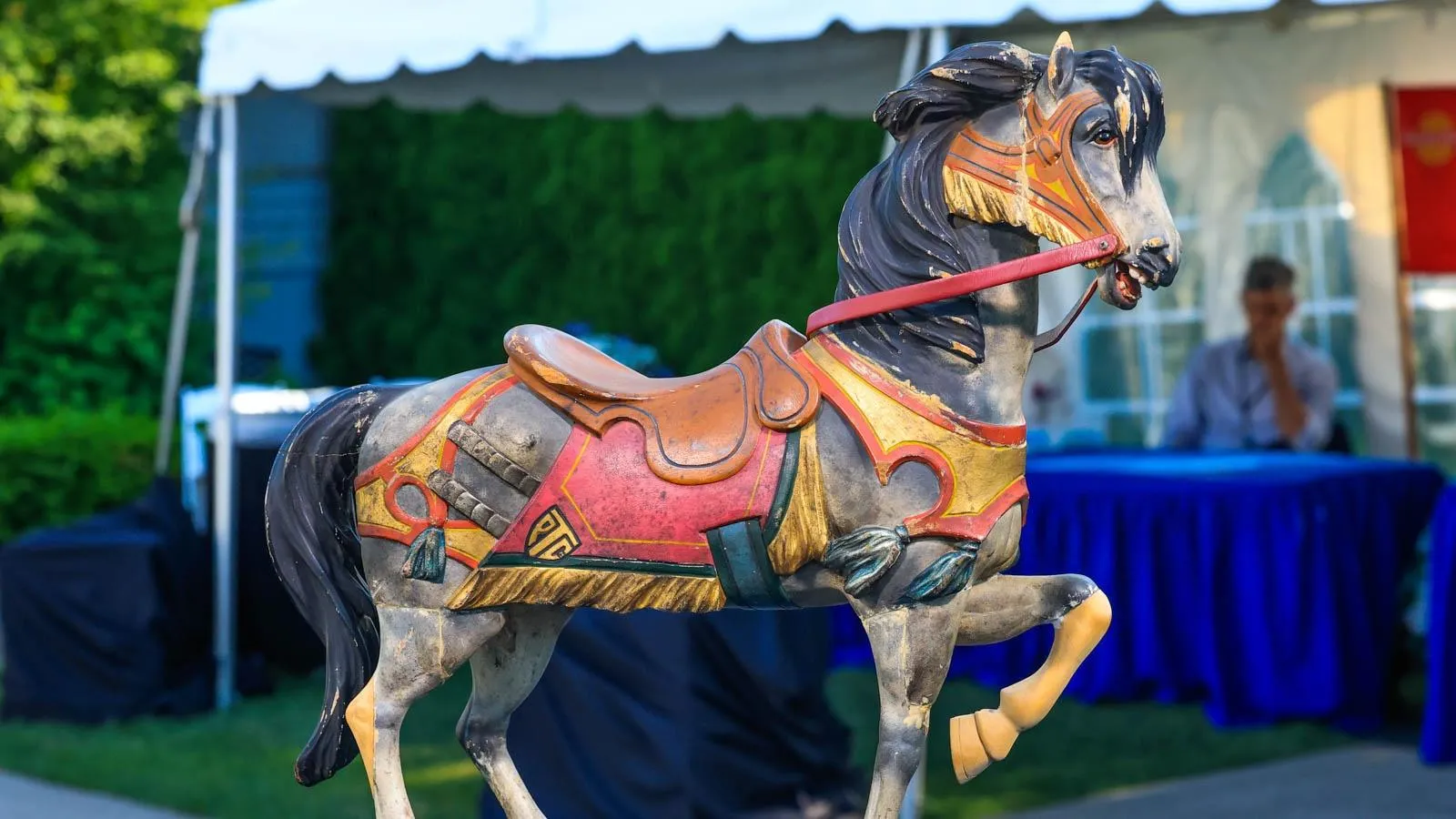A carousel horse with a red and gold saddle placed in front of an outdoor backdrop.