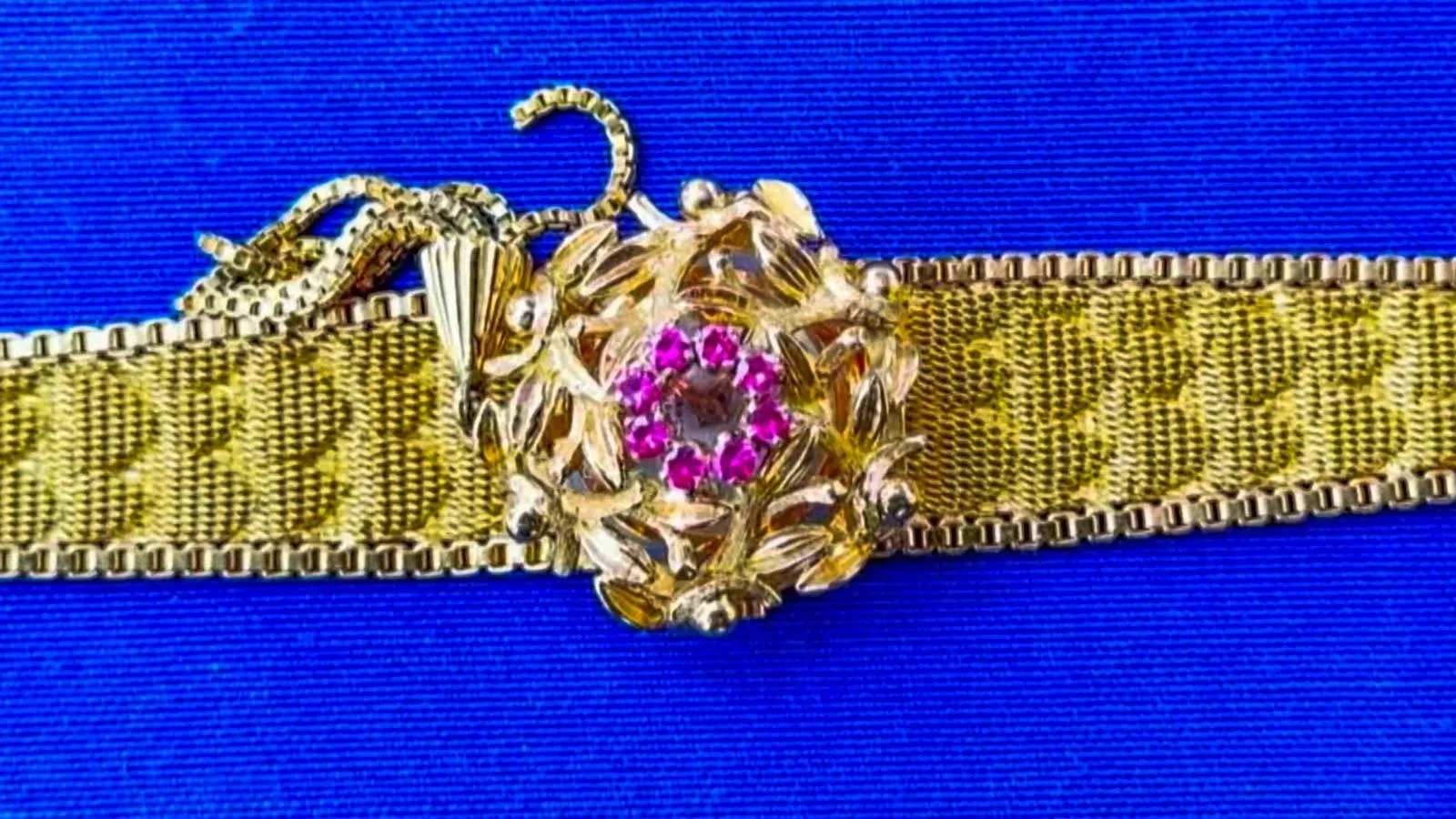 A yellow gold weaved bracelet with a center gold flower and pink sapphires in the middle against a blue background.