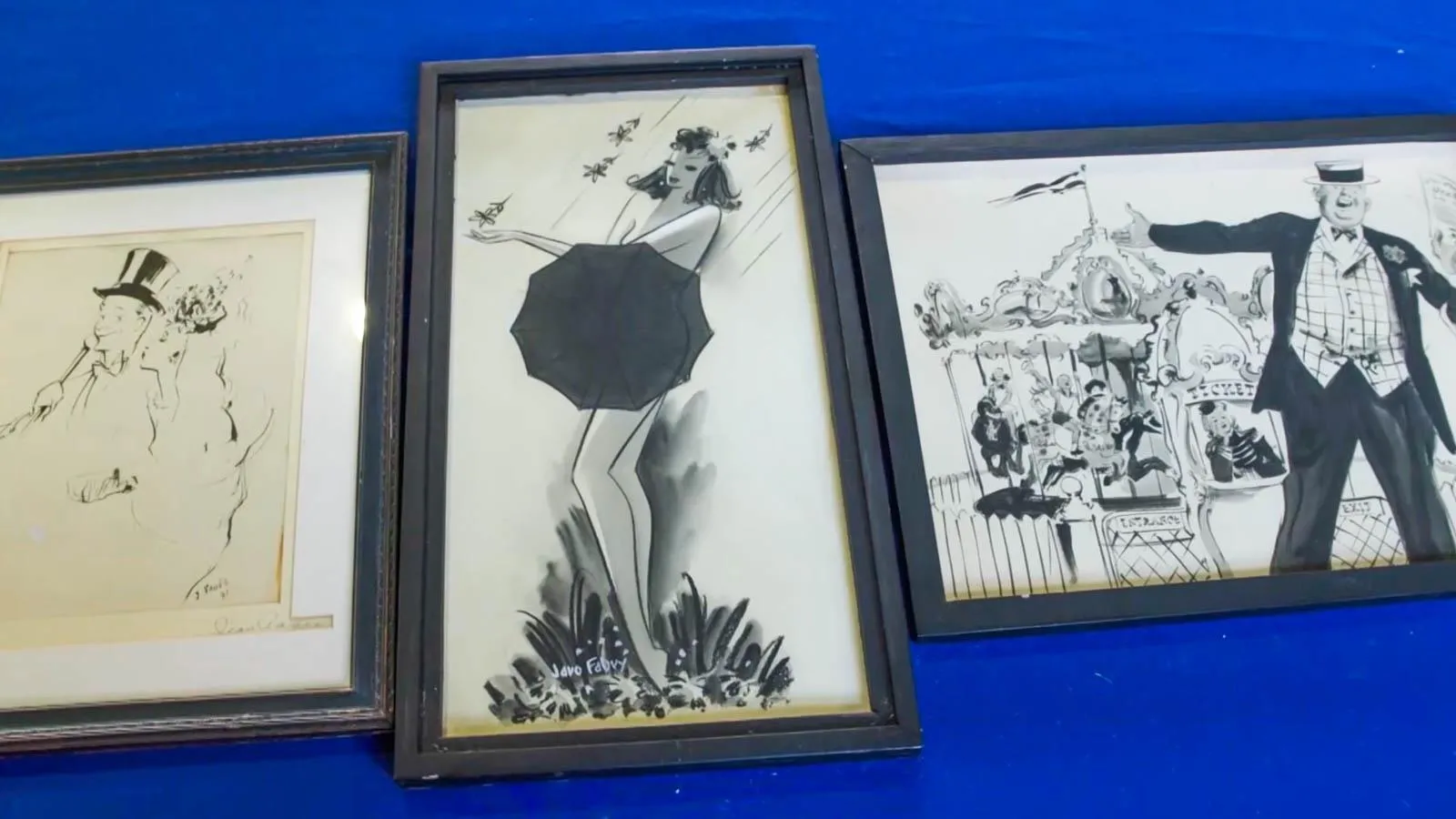 Three black and white drawings in black frames laid on top of a blue table. From left to right are drawings of a married couple, a female figure covered by an umbrella, and a man in a top hat.
