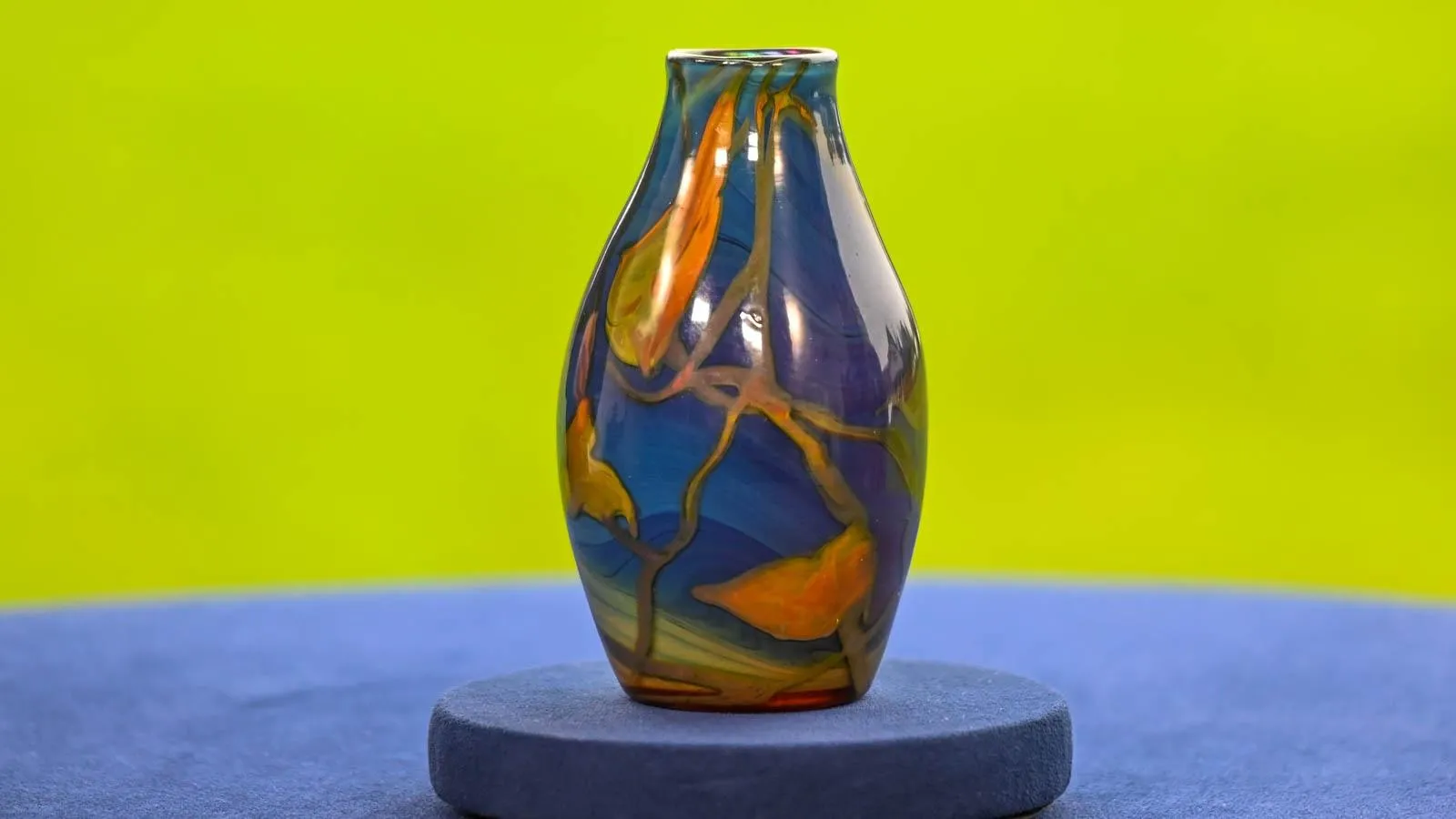 A small blue vase with orange accents sits on a table against a blue and green background.