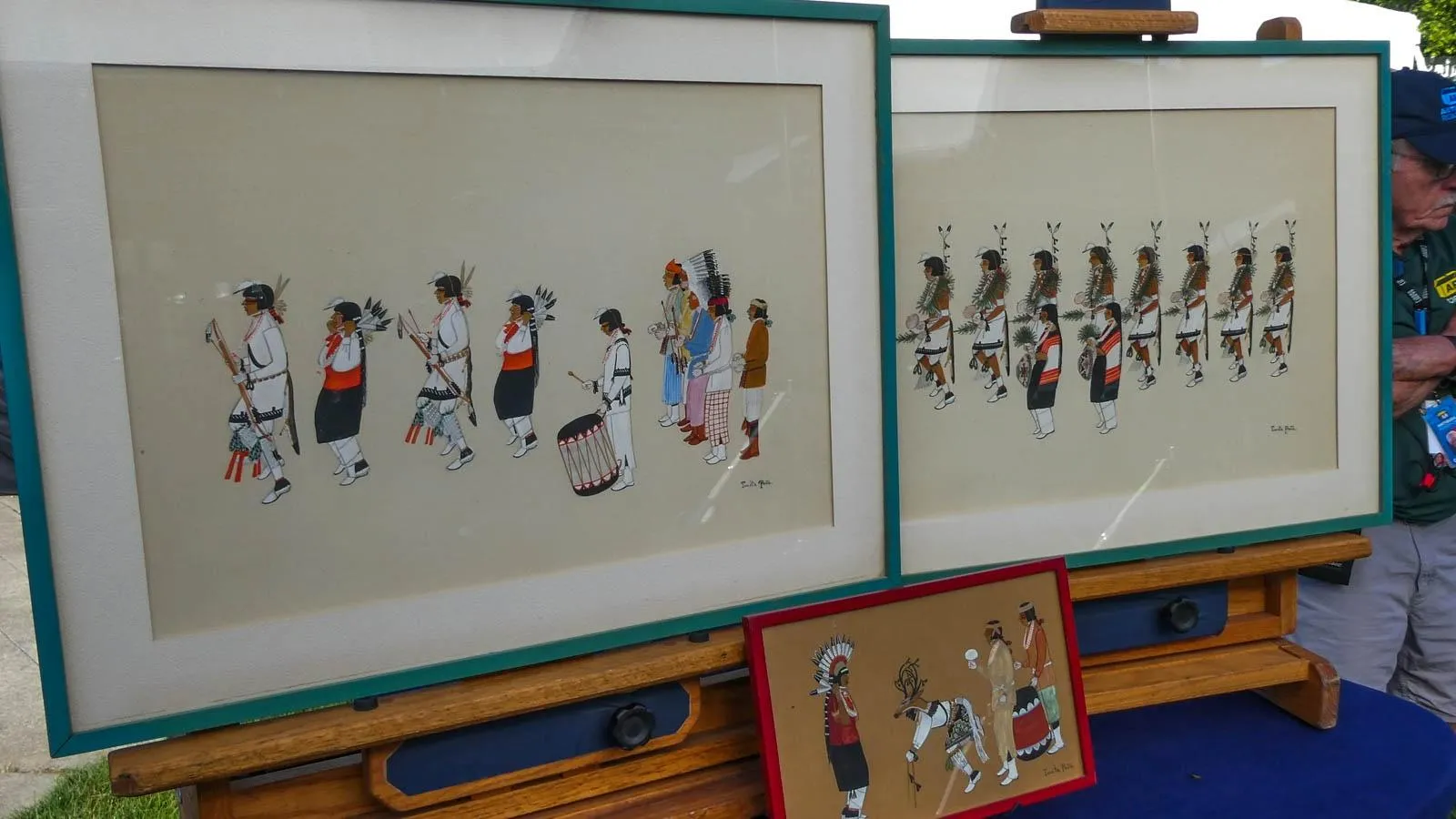 Two large paintings of people in a line are placed in a teal frame on top of a wooden shelf. Below these paintings is a smaller of people looking at an animal in a red frame placed on a blue table.