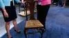 Two people standing on either side of a wooden chair with a yellowing floral cushion.