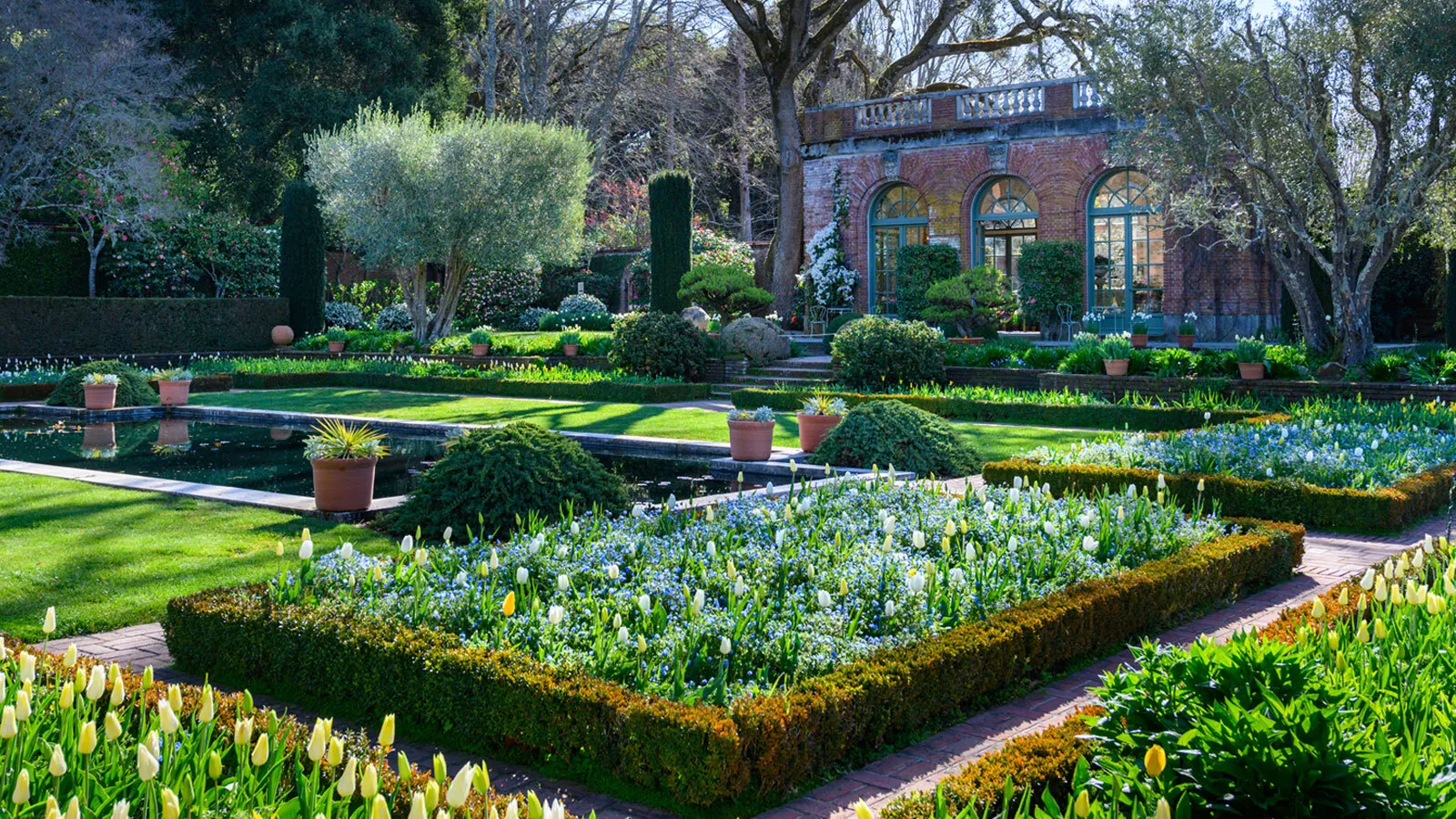 A formal garden of flower buds, with trees and shrubs surrounding each arrangement. A pond is situated near the center, across an antique brick structure with a terrace.