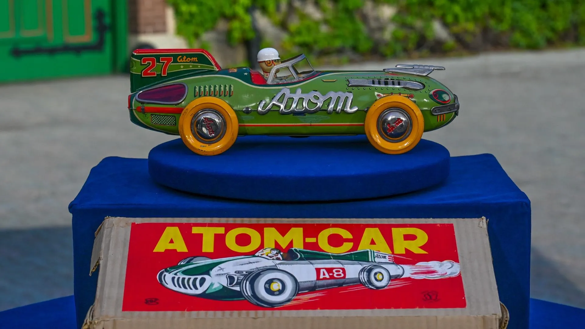 A tin green atom car placed on top of its box on a blue display plate.