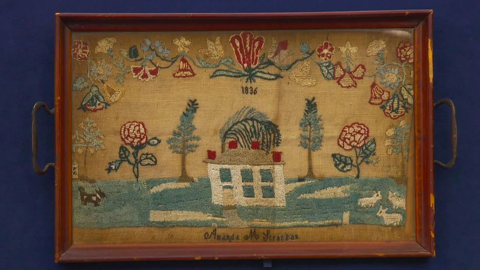 A framed sampler depicts a house in the middle of a green, flat landscape. Features small animals and floral designs that border the top of the illustration. Embroidery is done on brown burlap fabric.