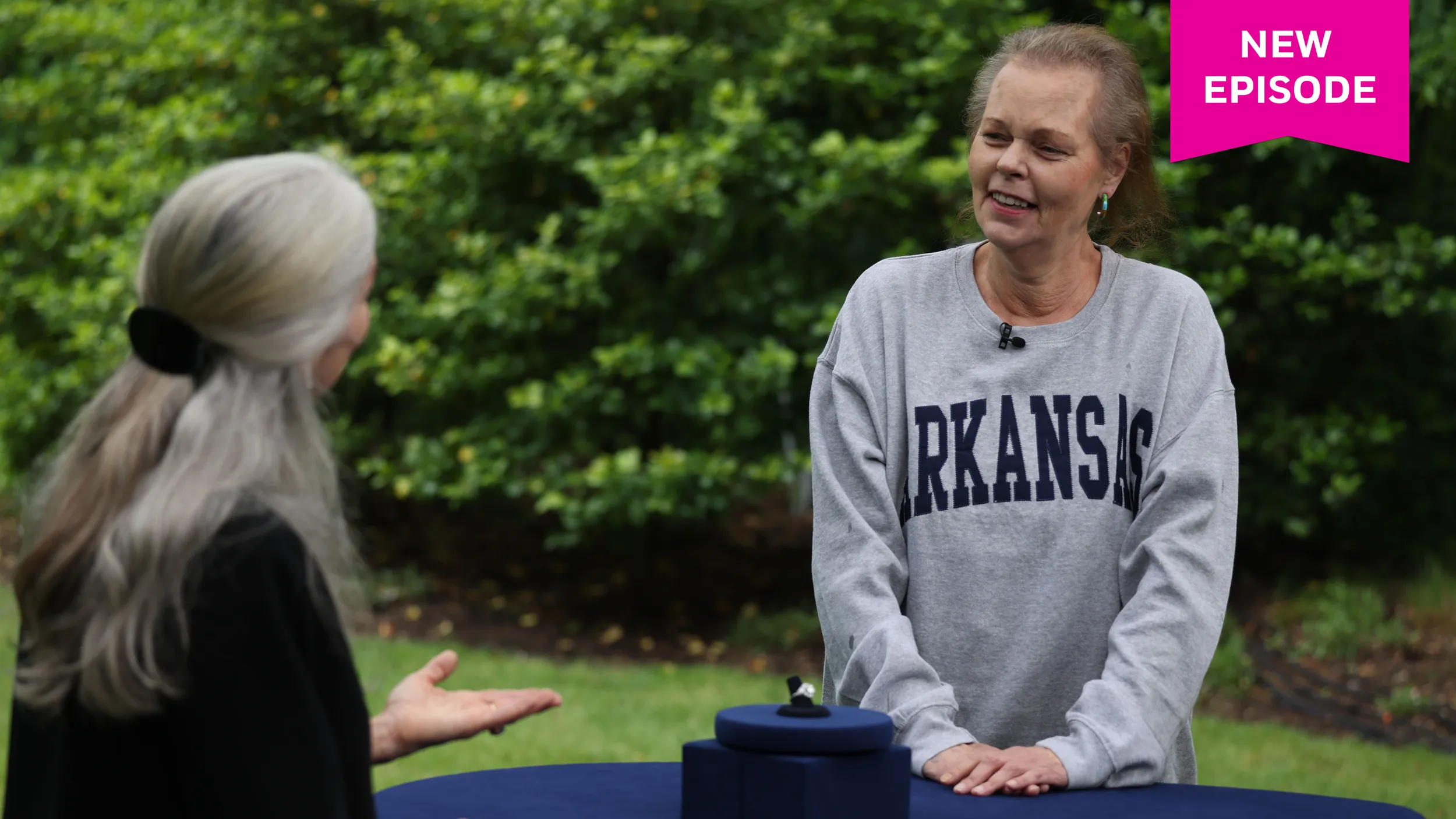 A woman in a sweatshirt that reads "Arkansas" looks shocked at an appraiser whose back is to the camera. On the table between them is a large diamond and platinum ring.