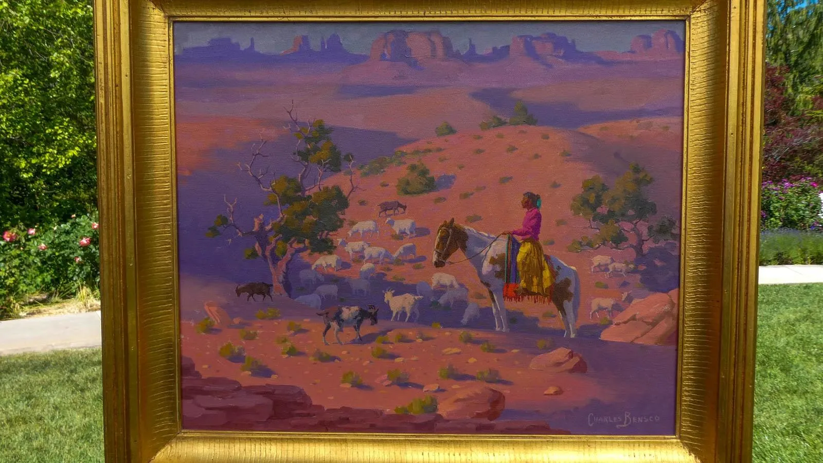 A desert scene of a Native American sitting on a white horse, guiding various sheep and goats down a rocky terrain in the desert mountains under the dusky sky.