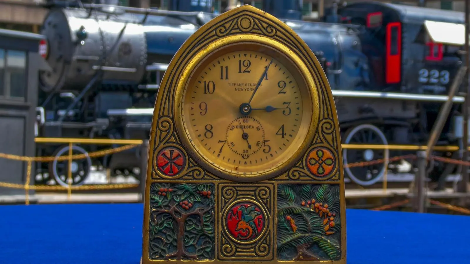 A clocked placed in a blue table with a train in the background. The clock is gold with green and orange nature themed decoration.