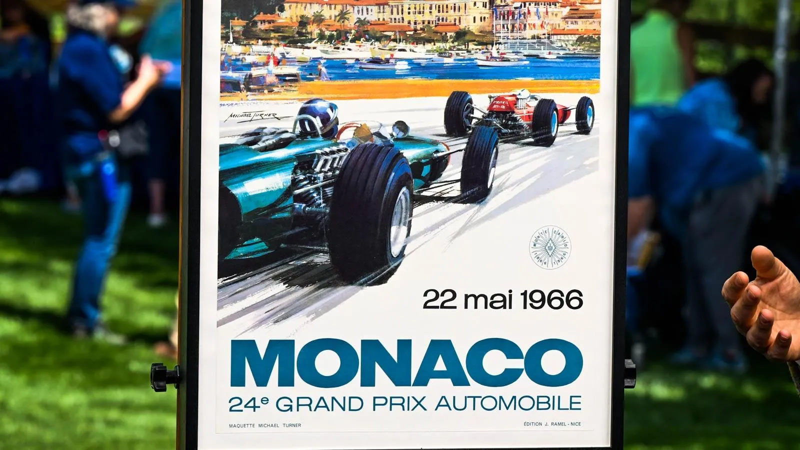 A poster with turquoise text in its lower portion that includes  “Monaco.” On the upper portion, there is a color illustration depicting race car drivers racing on a track that overlooks water.