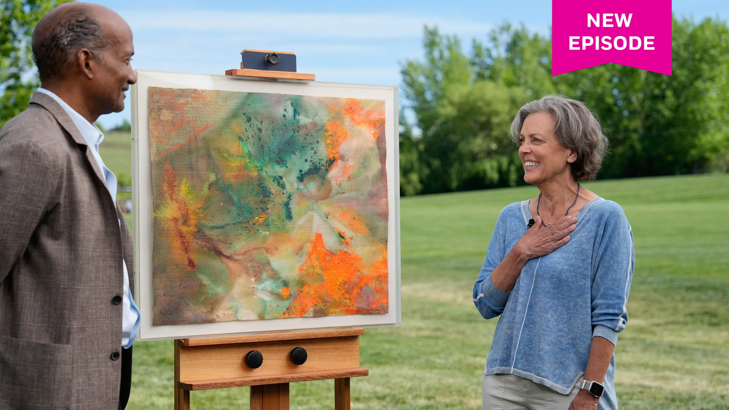 A woman in a blue shirt puts her hand over her heart as she smiles to an appraiser. Between them is an orange, green, and gray large abstract painting.