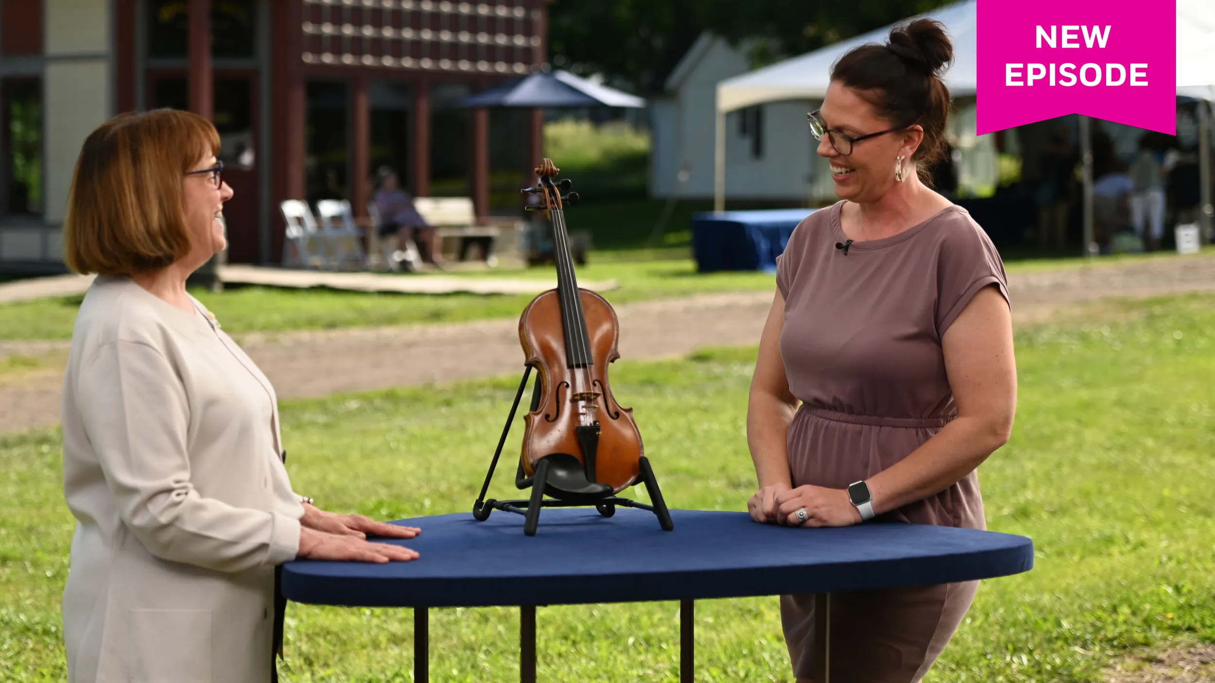A middle aged woman and a younger woman smile at each other. In between them is a small violin propped up on a blue appraisal table. They are on an outdoor film set with a white tent in the background