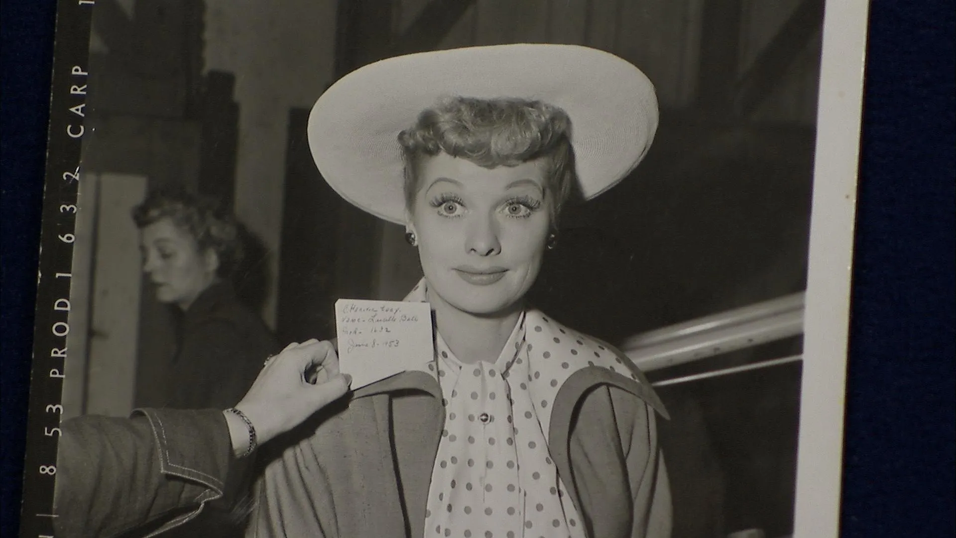 Close-up of a monochromatic photograph of actress "Lucille Ball" in costume, with a person holding a card to the left, which has written details and the date.