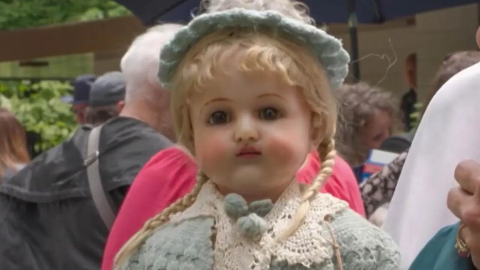 Close up of a doll with blonde braided hair, a blue bonnet, rosy cheeks, and blue petticoat.