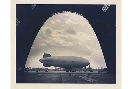 Black-and-white photo of a large oval aircraft parked on the tarmac at an airport. The photo is taken from inside a hanger looking out a tall archway towards the aircraft.